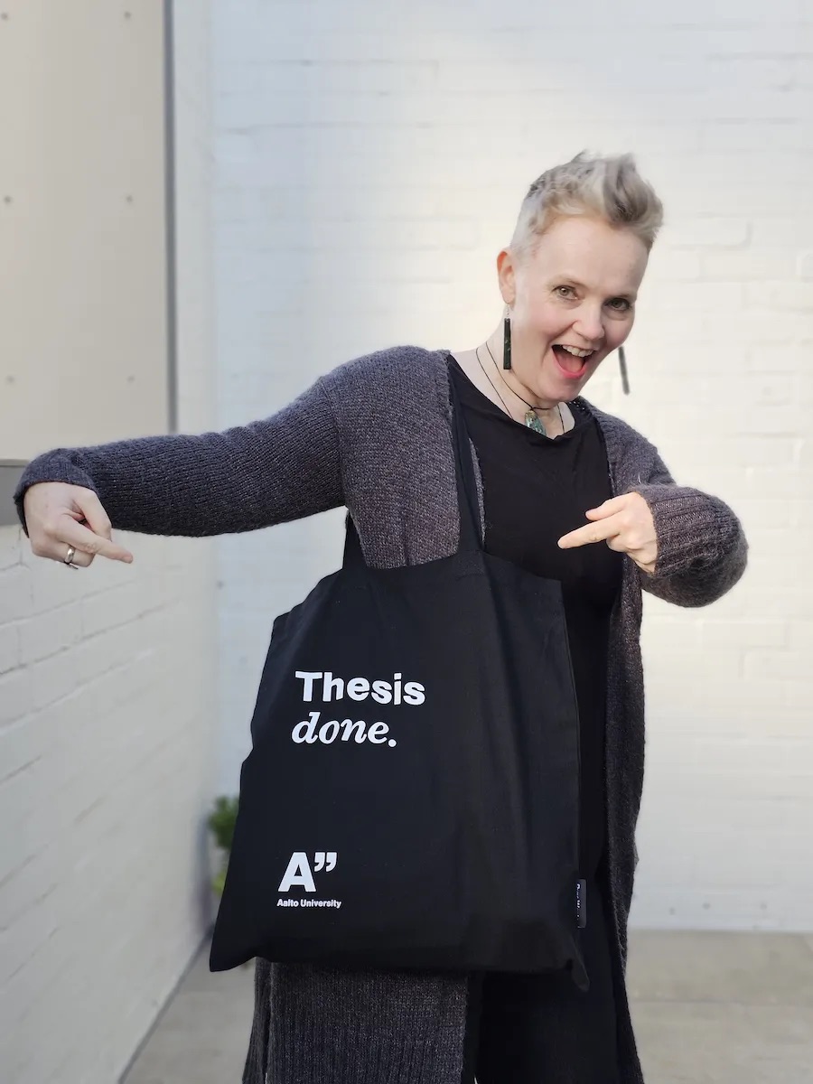 Photo of Liesel Mitchell smiling with a black tote bag with white writing saying 'thesis done'.